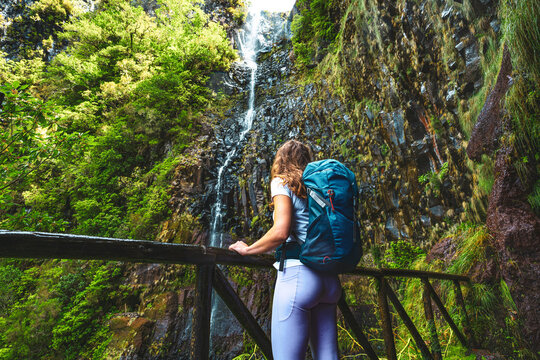 Sportive tourist with backpack leans against wooden fence of viewpoint and watches green overgrown Risco waterfall. 25 Fontes Waterfalls, Madeira Island, Portugal, Europe.