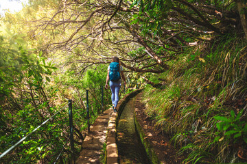 Female tourist hiking along a water canal path overgrown with trees on a sunny day. 25 Fontes Waterfalls, Madeira Island, Portugal, Europe.