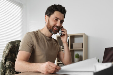 Happy soldier talking on phone at wooden table indoors. Military service