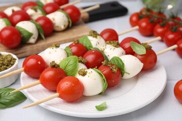 Caprese skewers with tomatoes, mozzarella balls, basil and pesto sauce on white table, closeup