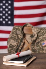 Notebooks and diploma on wooden table, chair with soldier uniform against flag of United States...