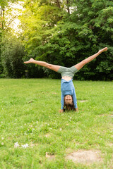 Child is engaged in acrobatics in the meadow at city park. The concept of a healthy lifestyle. Girl stands on her hands upside down, performs the wheel element. Fun and play on the grass in the park.