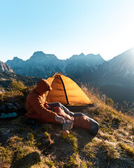 Camping in the wilderness. Man outside his tent is cooking breakfast during the sunrise in the mountains. © Luk