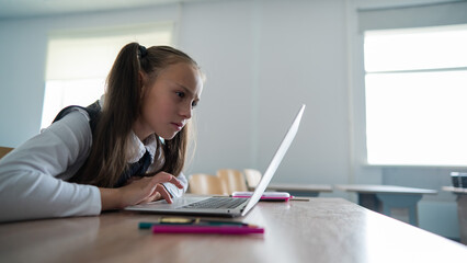 Caucasian girl sits at a desk at school and carefully looks into a laptop. 