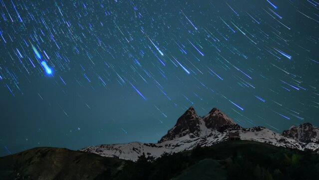 Mountain Ushba with peak covered by snow and sky with rotating stars and comets in timelapse. Night landscape of rock ridge under bright moving constellations and celestial bodies. Astronomy theme