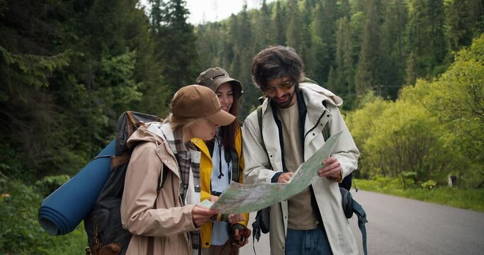 A trio of tourists get lost and are looking at a map to figure out where to go next. People in hiking clothes sort out where to have next in mountainous terrain