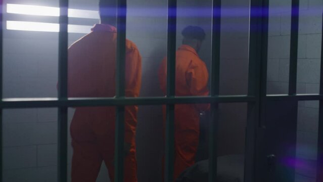 Two prisoners in orange uniforms emotionally talk in jail cell. Criminals start fighting. Jailer comes and puts on handcuffs on them. Inmates serve imprisonment terms for crimes in detention center.