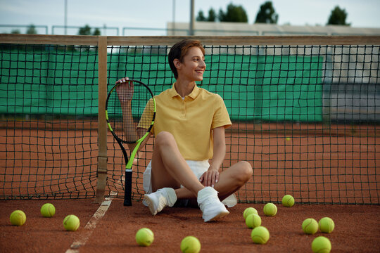 Attractive smiling woman tennis player sitting on court among scattered balls - Powered by Adobe