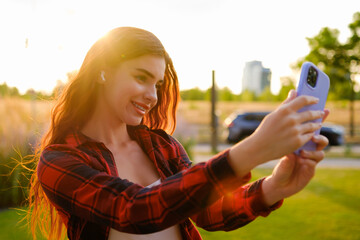 Red haired wearing red and black plaid shirt girl takes a selfie at sunset using a smartphone. 
