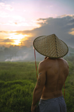 A young man wearing an Asian cone hat in rice fields at sunset. Bali.