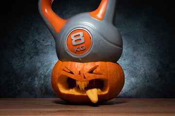 Heavy kettlebell crushing carved Halloween pumpkin head Jack Lantern (Jack-o'-lantern). Healthy diet, fitness lifestyle composition. Gym workout and sport training concept.