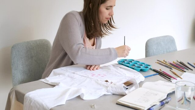 Caucasian woman painting a floral design in a white shirt. Unique clothing designs.