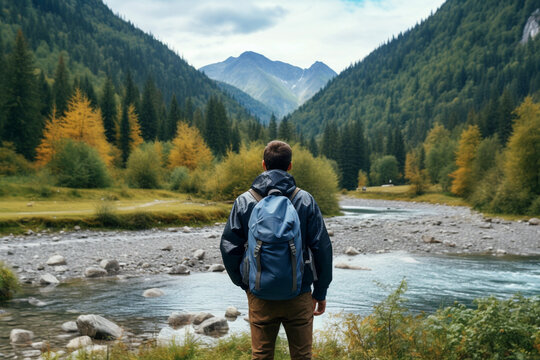 Unrecognizable adult with a blue backpack travels along the mouth of a mountain river against the backdrop of a wild forest, Rear View
