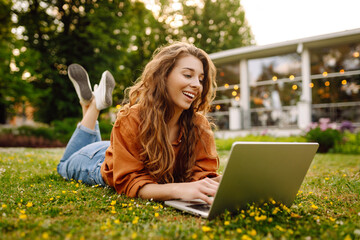 Portrait of a young beautiful female freelancer sitting on a green meadow with a laptop. Education online. Happy woman running outdoors.