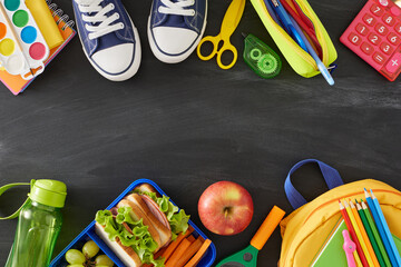 Balanced school break concept. Top view photo of lunchbox, organic treats, water bottle, assorted supplies, child's rucksack, footwear on blackboard background with empty space for promo or text