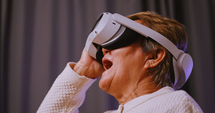 Asian old woman having fun playing games while wearing virtual reality glasses at home.