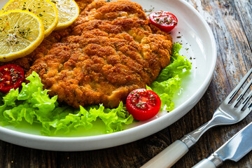 Crispy breaded seared chicken cutlet with lemon slices and fresh vegetables on wooden table
