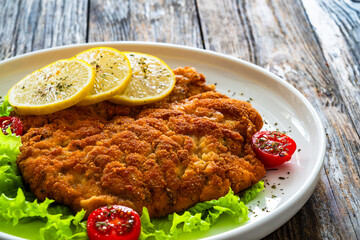 Crispy breaded seared chicken cutlet with lemon slices and fresh vegetables on wooden table
