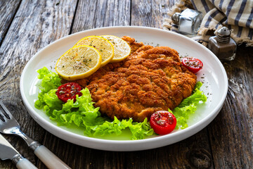 Crispy breaded seared chicken cutlet with lemon slices and fresh vegetables on wooden table
