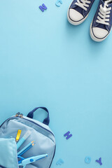 Education for young minds concept. Top view vertical shot of trendy sneakers, rucksack, pencil box, pencils, scissors, marker, blue letters on pastel blue background with empty space for promo or text