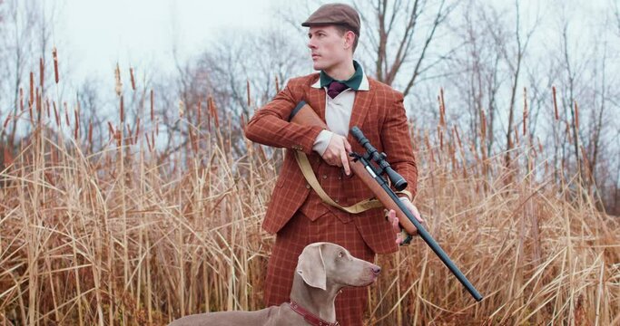 serious confident hunter in traditional shooting clothes outfit concentrated on watching birds, some game, standing on the field with dog, man is ready to hunt Slow motion, tradition