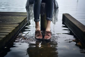 Poster Unrecognizable girl or young woman sitting on jetty by lake has taken off shoes and splashing bare feet in water © alisaaa