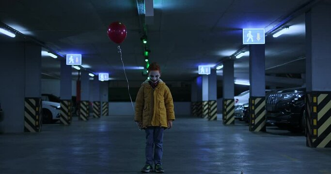 Cinematic shot of little boy in spooky scary clown make-up with balloon standing in a dark parking lot with flickering light and smiling ominously. Halloween costume party, horror movie concept
