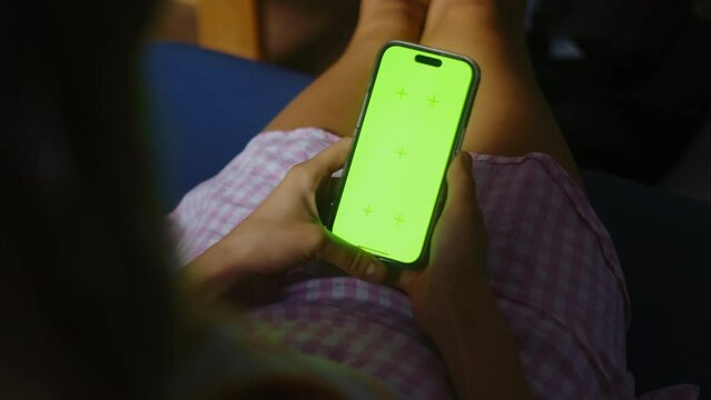 Young woman sitting on sofa at home and uses Mobile phone with green screen Mock-up. 