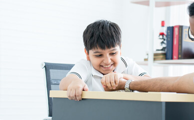 Naklejka premium Little handsome Indian child son and his father, have fun playing, competing in arm wrestling together on table at home. Cute boy win his dad. Education, Family relationship, Activity Concept
