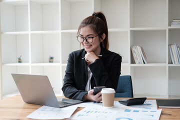 Smart Asian businesswoman working with laptop computer while holding smartphone in her hands, sitting at office desk.