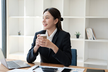 Young adorable Asian businesswoman holding a coffee cup while sitting at her office desk. taking a break from working.