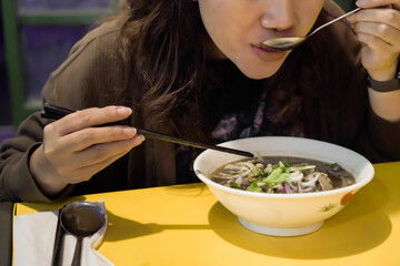 Asian woman eating Vietnamese soup dish Pho that consisting of broth, rice noodles, herbs, and beef...