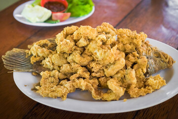 Ikan Gurame Goreng or fried freshwater fish with chilli sauce and vegetable. Serving on white plate on wooden table. Indonesian food and cuisine. Delicious Fried Gouramy.