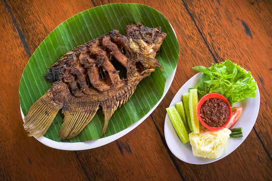 Ikan Gurame Bakar or grilled freshwater fish with chilli sauce and vegetable. Serving on white plate on wooden table. Indonesian food and cuisine. Delicious roasted Gouramy.