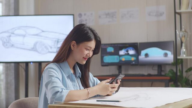 Asian Female Comparing Her Design To The Photo On Her Smartphone While Working On Car Design Sketch On Table In The Studio With Tv And Computers Display 3D Electric Car Model 
