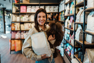 Beautiful young woman enjoying in modern pet shop together with her adorable brown toy poodle.  She is holding dog food bag.