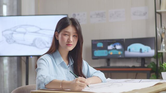 Asian Female Smiling To The Camera While Working On A Car Design Sketch On Table In The Studio With Tv And Computers Display 3D Electric Car Model 
