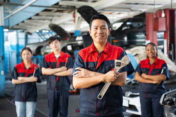 Selective focus of mid-adult Asian male mechanic in uniform, standing to hold a large adjustable spanner with arms folded and smiling at camera with a blurred mechanic team standing in the background.