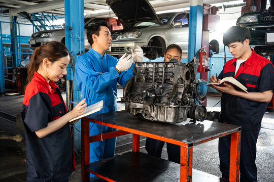 A middle-aged Asian senior male mechanic in blue uniform, standing to explain about the car engine placed on table to three junior mechanics who were listening and writing down in notebooks at garage.