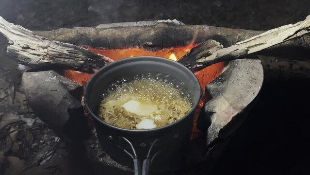 Boiling instant noodles and egg in a field pot on stove over a fire for cooking in a campsite. Bonfire cooking. Concept of travel and camping. Late night meal in camping.