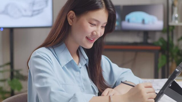Close Up Of Asian Female Smiling And Showing Thumbs Up Gesture To The Camera While Drawing New Car Design Concept On A Tablet In The Studio With Tv And Computers Display 3D Electric Car Model 
