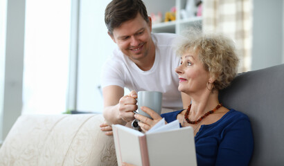 Fototapeta na wymiar Portrait of smiling man giving cup of tea to mom. Woman sitting on sofa and reading book. Family spending time together. Warm relationship concept