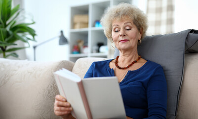 Portrait of adult woman reading interesting book. Aged curly-haired grandmother sitting on sofa at home. Smiling older grandma enjoying free time. Retirement vacation concept