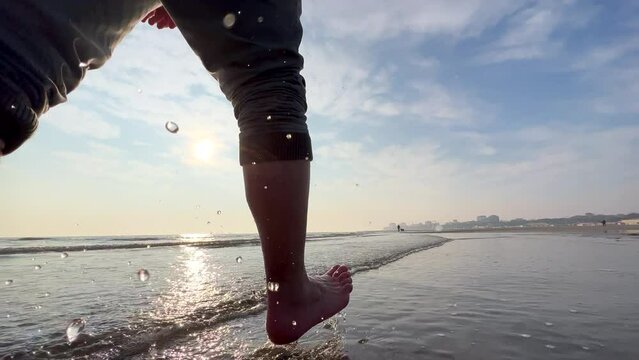 SLO MO Man walking on a sandy beach with coastal city at the background. Male feet splashing water. Seaside vacation