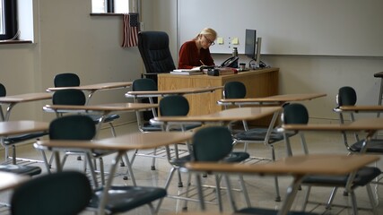Happy woman teacher sitting at desk in empty school classroom desk grading papers working on assignments. US American flag in the background.