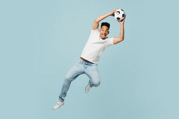 Full body goalkeeper happy fun young man fan wearing t-shirt cheer up support football sport team hold catch soccer ball jump high watch tv live stream isolated on plain pastel blue color background.