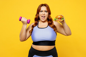Young angry chubby overweight plus size big fat fit woman wear blue top warm up training hold in hand burger and dumbbells isolated on plain yellow background studio home gym. Workout sport concept.