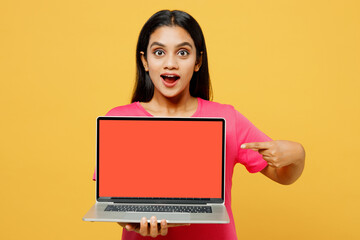Naklejka premium Young surprised IT Indian woman wearing pink t-shirt casual clothes hold use work point on laptop pc computer with blank screen workspace area isolated on plain yellow background. Lifestyle concept.