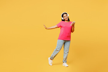 Fototapeta na wymiar Full body young happy Indian woman wearing pink t-shirt casual clothes use mobile cell phone listen to music in headphones dance isolated on plain yellow background studio portrait. Lifestyle concept.