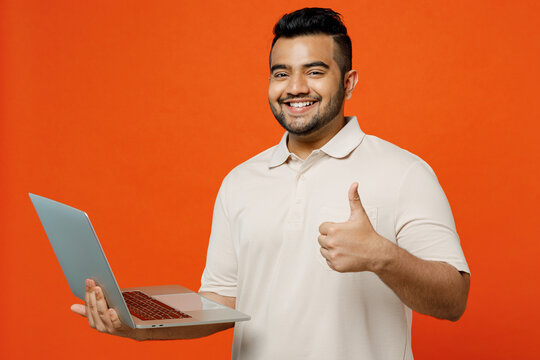 Young fun smiling smart IT Indian man he wears white t-shirt casual clothes hold use work on laptop pc computer show thumb up isolated on plain orange red background studio portrait Lifestyle concept